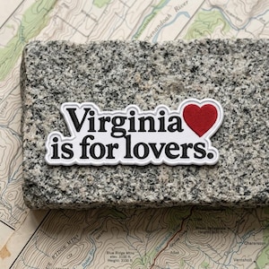 May include: A rectangular embroidered patch with a white background and a black border. The text reads "Virginia is for lovers." in black, bold, serif lettering, with a solid red heart icon replacing the letter "o" in the word "lovers."