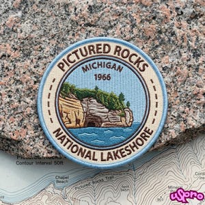 May include: A circular embroidered patch featuring a landscape of Pictured Rocks, Michigan, with the text Pictured Rocks National Lakeshore and 1966. The design includes tan cliffs, green trees, and blue water, all bordered by a light blue stitched edge.