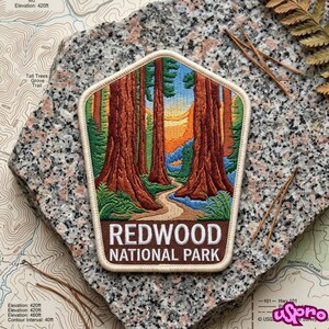 May include: A pentagonal embroidered patch featuring a landscape of tall redwood trees, a winding path, and a sunset sky. The bottom brown border contains the white text REDWOOD NATIONAL PARK. The patch has a cream-colored stitched border edge.