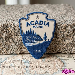 May include: A shield-shaped embroidered patch featuring a cream-colored background with dark blue text reading ACADIA MAINE and the year 1919. The design includes a dark blue embroidered landscape of pine trees and a small star at the top.