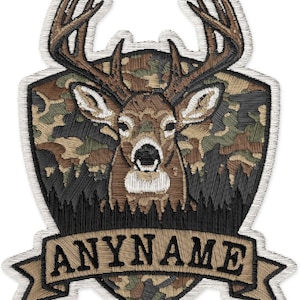 May include: An embroidered patch featuring a deer head with antlers centered on a shield-shaped background with a camouflage pattern. Below the deer, a tan banner displays the text ANYNAME in black capital letters, all outlined in white.