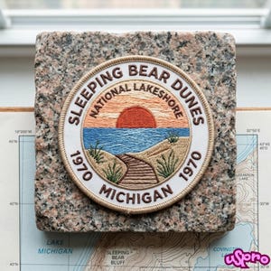 May include: A circular embroidered patch featuring a landscape of Sleeping Bear Dunes National Lakeshore. The design includes a red sun, blue water, a wooden path, and green grass. Text reads: SLEEPING BEAR DUNES NATIONAL LAKESHORE 1970 MICHIGAN.