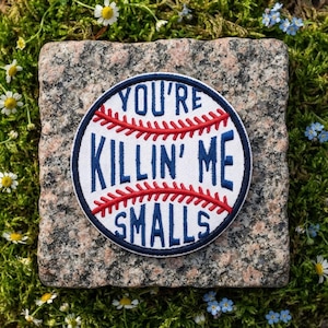 May include: A circular embroidered patch featuring a white baseball design with red stitching and dark blue text that reads YOU'RE KILLIN' ME SMALLS. The patch has a dark blue border and is centered on a square piece of speckled granite stone.