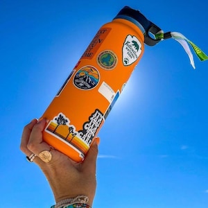 K&ouml;nnte beinhalten: Eine orangefarbene, isolierte Wasserflasche mit mehreren Aufklebern, darunter Yellowstone National Park und ein Sonnenuntergangsdesign. Die Flasche hat einen schwarzen Deckel und einen Griff mit einem gr&uuml;n-wei&szlig;en Anh&auml;nger. Der Hintergrund ist ein strahlend blauer Himmel.