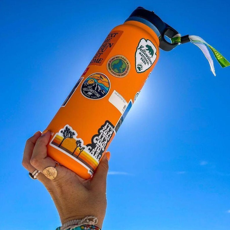 K&ouml;nnte beinhalten: Eine orangefarbene, isolierte Wasserflasche mit mehreren Aufklebern, darunter Yellowstone National Park und ein Sonnenuntergangsdesign. Die Flasche hat einen schwarzen Deckel und einen Griff mit einem gr&uuml;n-wei&szlig;en Anh&auml;nger. Der Hintergrund ist ein strahlend blauer Himmel.