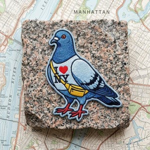 May include: An embroidered patch featuring a blue and grey pigeon wearing a white shirt with a red heart and the text I NY, along with a yellow crossbody bag. The pigeon has dark blue wing feathers and bright red feet, rendered in detailed thread.