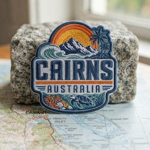 May include: An embroidered patch featuring a sunset over mountains, a wave, a palm tree, and a sea turtle. The text reads CAIRNS AUSTRALIA in bold blue letters. The design includes orange, yellow, white, and various shades of blue thread.