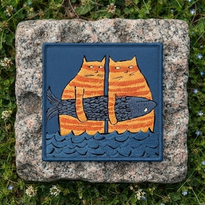 May include: A square embroidered patch featuring two orange tabby cats with dark stripes standing in dark blue water. The cats are holding a large, dark gray fish horizontally between them. The background of the patch is a solid dark blue color.