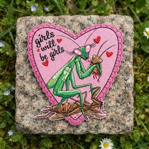 May include: A heart-shaped embroidered patch with a pink background featuring a green praying mantis eating a brown insect. The text "girls will be girls" is stitched in black, accented by small red hearts surrounding the central illustration.