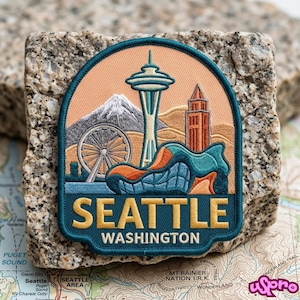 May include: A circular embroidered patch featuring a Seattle cityscape with the Space Needle, a Ferris wheel, and a mountain peak against a peach sky. The lower section is dark teal with the yellow text SEATTLE and white text WASHINGTON.