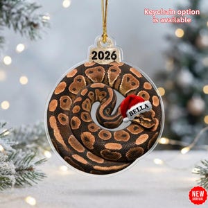 May include: A clear, circular acrylic ornament featuring a coiled brown and black patterned snake wearing a small red and white Santa hat with the name BELLA printed on the brim. The year 2026 is printed in black at the top, and a gold string is attached.