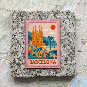 May include: A rectangular embroidered patch featuring a stylized illustration of the Sagrada Familia in Barcelona, Spain. The design includes a bright orange sun, palm trees, and a colorful mosaic wall. The word BARCELONA is stitched in orange.