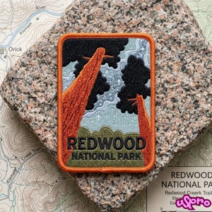 May include: A rectangular embroidered patch with an orange border featuring a landscape of two tall orange redwood trees, black foliage, and a light blue starry sky. The text REDWOOD NATIONAL PARK is embroidered in black at the bottom of the patch.