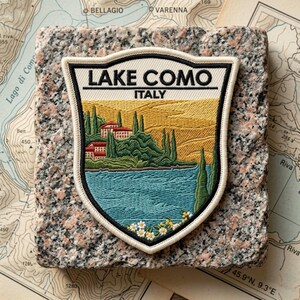 May include: A shield-shaped embroidered patch featuring the text LAKE COMO ITALY. The design depicts a blue lake, green cypress trees, red-roofed buildings, and yellow hills. The patch is centered on a square piece of speckled pink and gray granite.