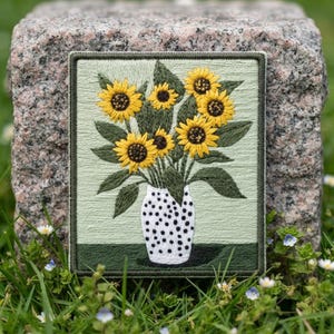 May include: A rectangular embroidered patch featuring a bouquet of yellow sunflowers with dark brown centers and green leaves arranged in a white vase with black polka dots, set against a light green background with a dark green base border.