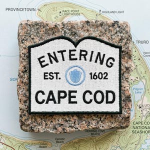 May include: A white embroidered patch with a black border and black text that reads ENTERING EST. 1602 CAPE COD, featuring a small light blue circular emblem in the center, resting on a square piece of speckled pink and grey granite stone.