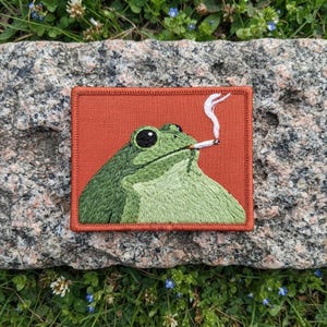 May include: A rectangular embroidered patch featuring a green frog with large black eyes on a solid burnt orange background. The frog is depicted in profile, holding a white cigarette in its mouth with a thin trail of white smoke rising upward.