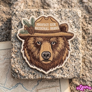 May include: An embroidered patch featuring a brown bear wearing a tan ranger hat with a pine branch and cone. The hat displays the text PROTECT OUR NATIONAL PARKS in white capital letters. The patch has a cream-colored border and detailed fur.