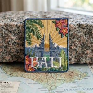 May include: A rectangular embroidered patch featuring a blue Balinese temple gate, yellow sunburst rays, green palm fronds, and pink tropical flowers. The word BALI is stitched in large white capital letters across the center of the design.
