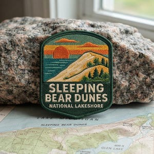 May include: A shield-shaped embroidered patch featuring a landscape of Sleeping Bear Dunes National Lakeshore. The design shows a sunset over water, a large sand dune, and green trees. The text reads SLEEPING BEAR DUNES NATIONAL LAKESHORE.