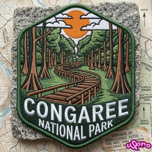 May include: A hexagonal embroidered patch featuring a brown boardwalk winding through a green forest with tall trees and a bright orange sun in the sky. The text CONGAREE NATIONAL PARK is stitched in white capital letters at the bottom of the patch.