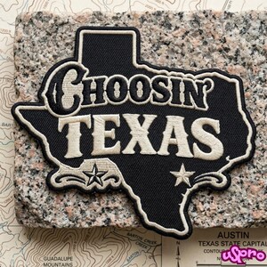 May include: A black embroidered patch shaped like the state of Texas with a cream-colored border. The text "CHOOSIN'" is stitched in black and "TEXAS" in cream. Two cream-colored stars are featured at the bottom of the patch design.