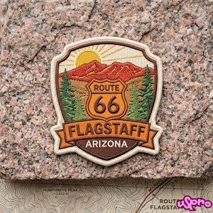May include: An embroidered patch featuring a landscape with a yellow sun, orange mountains, and green pine trees. A central shield displays the text ROUTE 66, with the words FLAGSTAFF and ARIZONA printed on banners below in dark brown and orange.