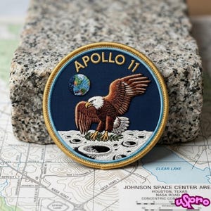May include: A circular embroidered patch featuring the Apollo 11 mission emblem. The design includes a brown and white eagle landing on the moon surface, a small Earth in the background, and the text APOLLO 11 in gold against a dark blue field.