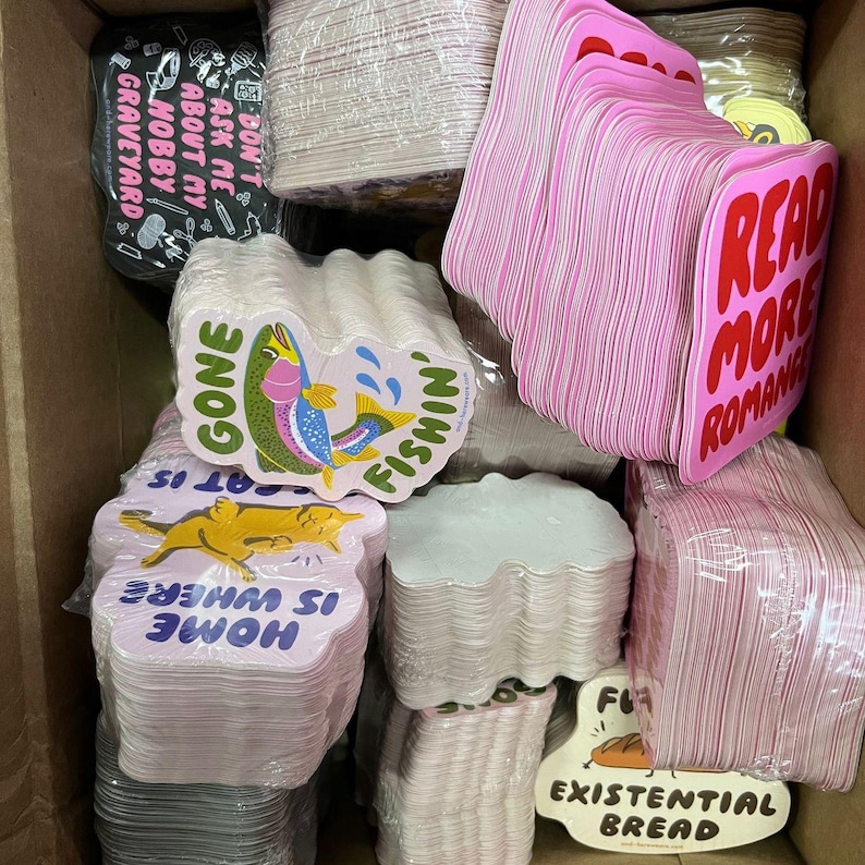 K&ouml;nnte beinhalten: Eine Kiste voller verschiedener Aufkleber. Einige Aufkleber sind rosa mit dem Text "READ MORE ROMANCE" in Rot. Andere Aufkleber haben S&auml;tze wie "GONE FISHIN'" und "HOME IS WHERE AT IS".