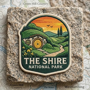 May include: An embroidered patch featuring a landscape with a round yellow door set into a green hill, a winding path, and a sunset sky. The text reads THE SHIRE NATIONAL PARK in white capital letters against a dark green background border.