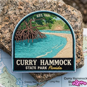 May include: A semi-circular embroidered patch featuring a landscape of a beach with mangrove trees, a sunrise, and the text EST. 1991, CURRY HAMMOCK STATE PARK Florida. The patch has a dark teal border and a black base with white and yellow lettering.