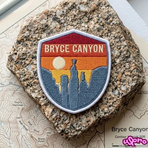 May include: A shield-shaped embroidered patch featuring a landscape of Bryce Canyon with a red, orange, and yellow sunset sky, a cream-colored sun, and blue rock formations. The text BRYCE CANYON is stitched in cream letters across the top.