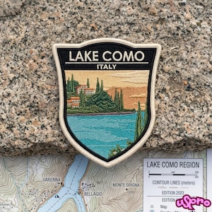 May include: A shield-shaped embroidered patch featuring a landscape of Lake Como, Italy. The design includes a blue lake, green trees, and buildings under a tan sky. The text LAKE COMO and ITALY is stitched in white on a black background.