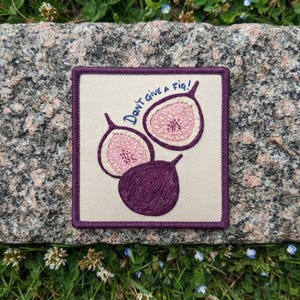 May include: A square embroidered patch on a cream fabric base featuring two halved figs and one whole fig in shades of purple and pink. The text DON'T GIVE A FIG! is stitched in blue thread above the fruit. The patch has a dark purple border.