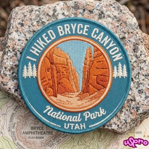 May include: A circular embroidered patch featuring a landscape of orange rock formations under a light blue sky. The text reads I HIKED BRYCE CANYON National Park UTAH. The design includes white pine tree silhouettes on a teal background with a border.