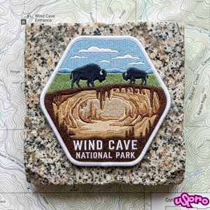 May include: A hexagonal embroidered patch featuring a landscape with two black bison standing on green grass under a blue sky with white clouds, above a brown cave interior with stalactites, with the text WIND CAVE NATIONAL PARK in white.