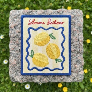 May include: A rectangular embroidered patch featuring a light yellow background with subtle vertical stripes. The text Limone Siciliano is stitched in red above three yellow lemons with green leaves, all enclosed by a wavy blue border.