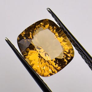 以下が含まれることがあります： クッションカットが施されたゴールデンイエローのシトリンの宝石。黒い金属製のピンセットで保持されています。宝石は透明度が高く、光を反射する鋭い幾何学的なファセットが特徴です。