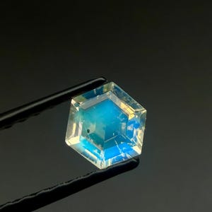 Può includere: Una gemma esagonale sfaccettata, con una vibrante gradazione di colore blu e giallo. Tenuta da pinzette nere su sfondo scuro, che ne evidenzia la chiarezza e la brillantezza. Le sfaccettature della gemma riflettono la luce, creando un'esposizione accattivante.