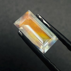 May include: A rectangular, step-cut gemstone featuring a translucent, colorless outer border and a vibrant, warm amber-orange core. The stone displays internal facets and light refraction, showcasing a clear transition from the pale edges to the center.