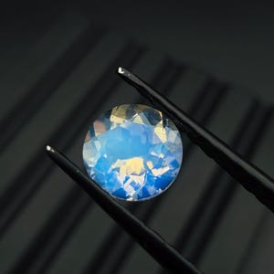Könnte beinhalten: Ein runder, facettierter Edelstein mit blau-gelbem Farbschema wird von einer schwarzen Pinzette vor einem dunklen Hintergrund gehalten. Die Facetten des Edelsteins reflektieren das Licht und erzeugen einen funkelnden Effekt.