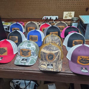 May include: A collection of trucker hats with various color combinations, including red, white, black, blue, and camouflage. Each hat features a leather patch with a design and text. The hats are displayed on a wooden surface.