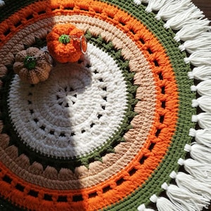 May include: A circular crochet mat featuring concentric rings of white, beige, orange, and dark green yarn, finished with a white fringe border. Two small, stuffed crochet pumpkin keychains in orange and beige sit on the mat surface.