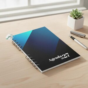 May include: A spiral-bound notebook with a gradient cover transitioning from light blue to dark blue and black. The cover features the white text Agenda 2027. A silver metal pen and a small potted succulent are positioned nearby on the surface.