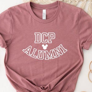 May include: A dusty rose t-shirt with the text "DCP ALUMNI" in white, arched across the front, with a Mickey Mouse head icon. The shirt has rolled-up sleeves and a knotted hem.