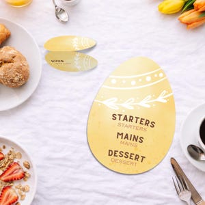 May include: A yellow egg-shaped menu card with the words "Starters", "Mains", and "Dessert". Two smaller oval cards are also visible. The cards have a white floral design and the text "HUUB VAN LIEN".