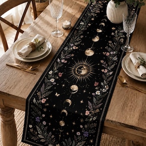 May include: A black rectangular table runner featuring a central sun illustration surrounded by a vertical sequence of moon phases, accented with botanical floral patterns, small stars, and butterflies in gold, white, and muted purple tones.