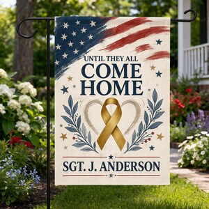 May include: A garden flag with a design featuring the American flag in red, white, and blue. The flag has the text "UNTIL THEY ALL COME HOME" and "SGT. J. ANDERSON". A gold ribbon and heart design is in the centre.