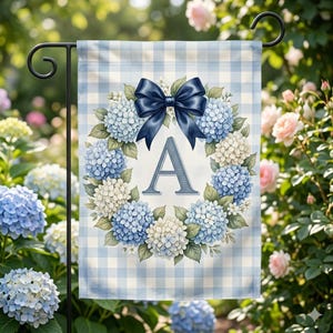 May include: A garden flag featuring a blue and white gingham pattern. The flag displays a wreath of blue and white hydrangeas, a navy blue bow, and the letter "A". The flag is hanging on a black metal stand.