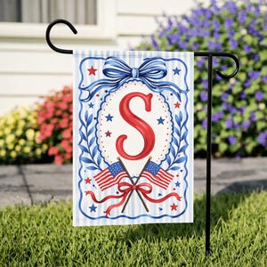 May include: A patriotic garden flag featuring a large red letter "S" within a white oval, surrounded by blue and white decorative elements. The flag includes crossed American flags and a blue bow at the top, set against a striped background.
